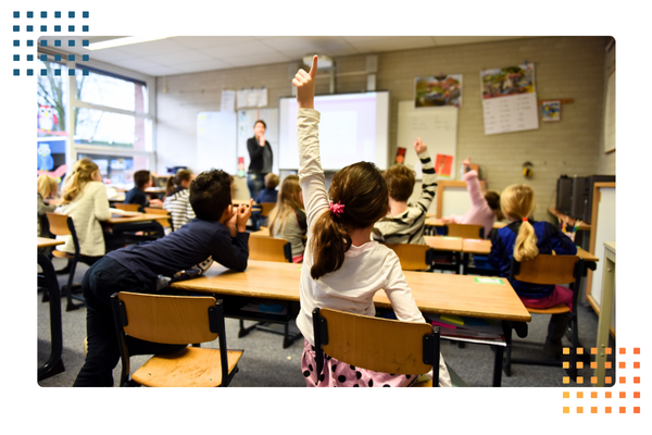 young girl raises her hand in the middle of an elementary school classroom while teachers calls on her