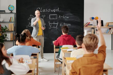 male teacher in front of a chalkboard with math problems calls on a student whose hand is raised